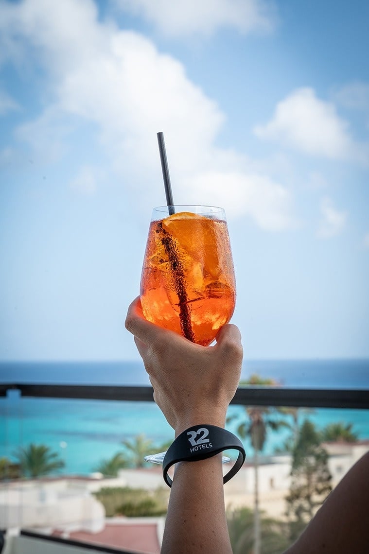 Hand holding a refreshing orange cocktail with an R2 Hotels wristband against a beautiful ocean view.