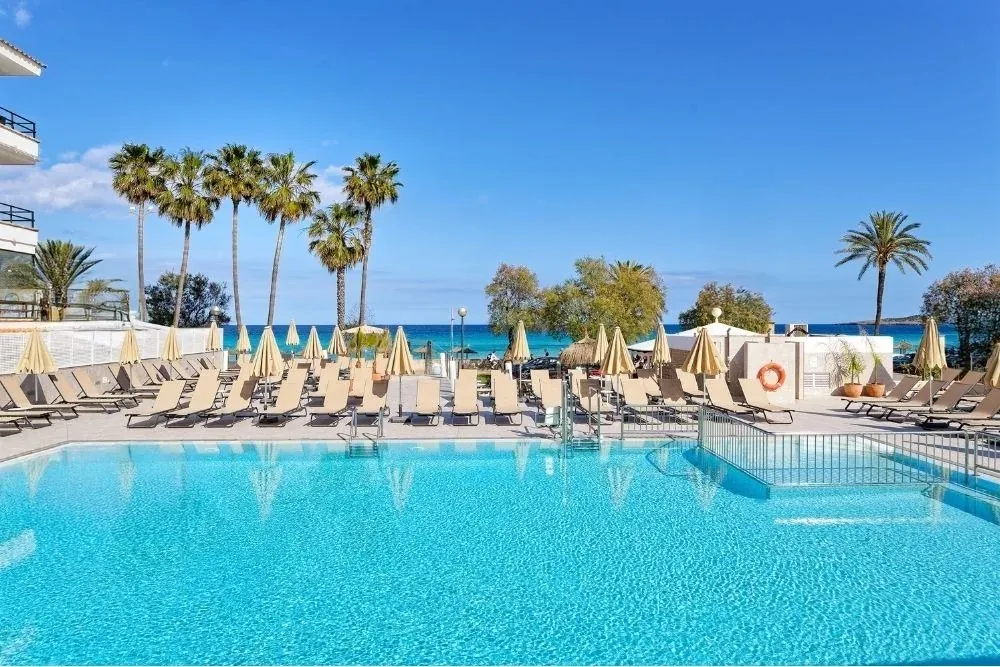 R2 Hoteles: Piscina azul frente al mar, tumbonas y palmeras bajo el sol.