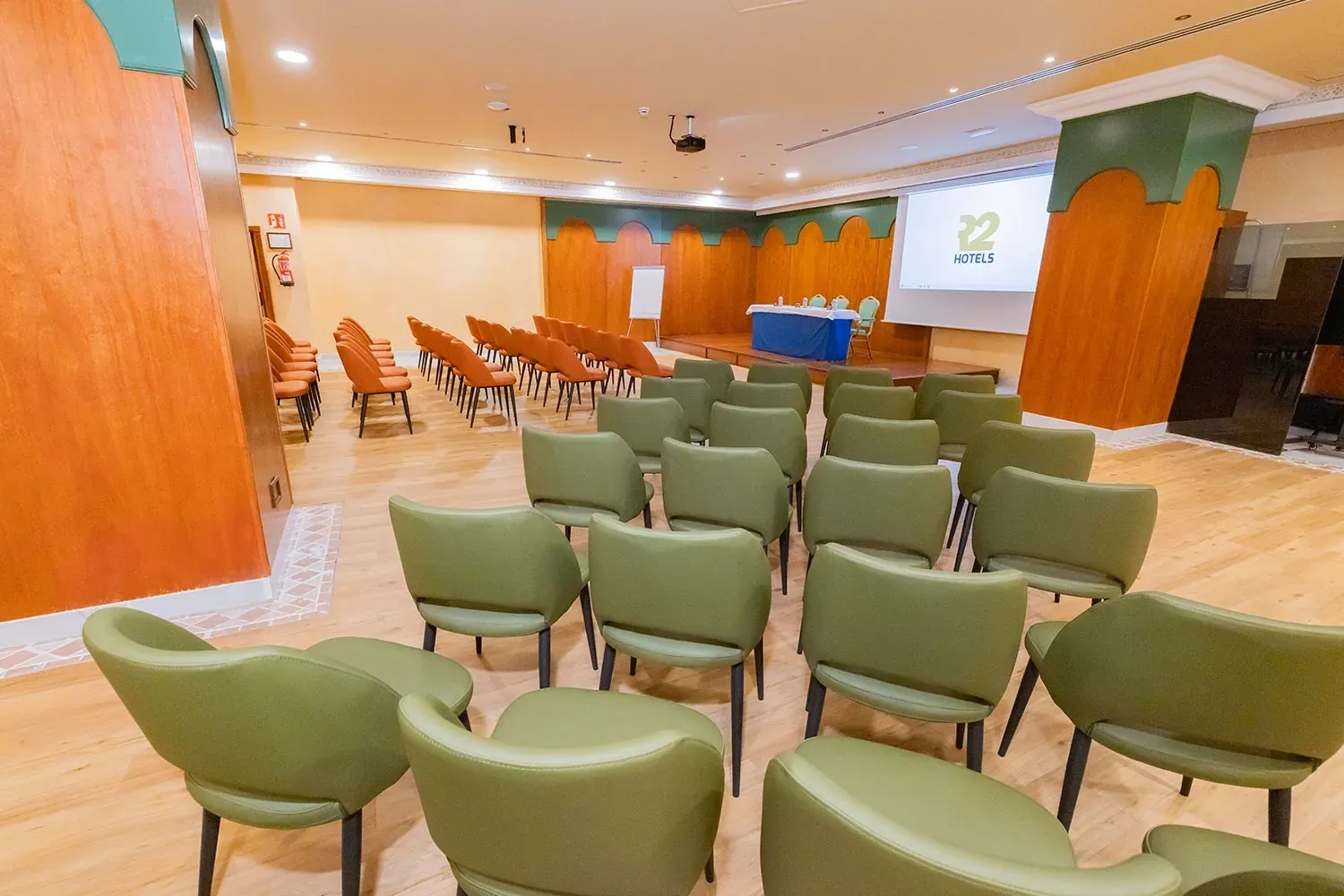 R2 Hotels Konferenzraum: grüne und orange Stühle, Bühne, Projektor.