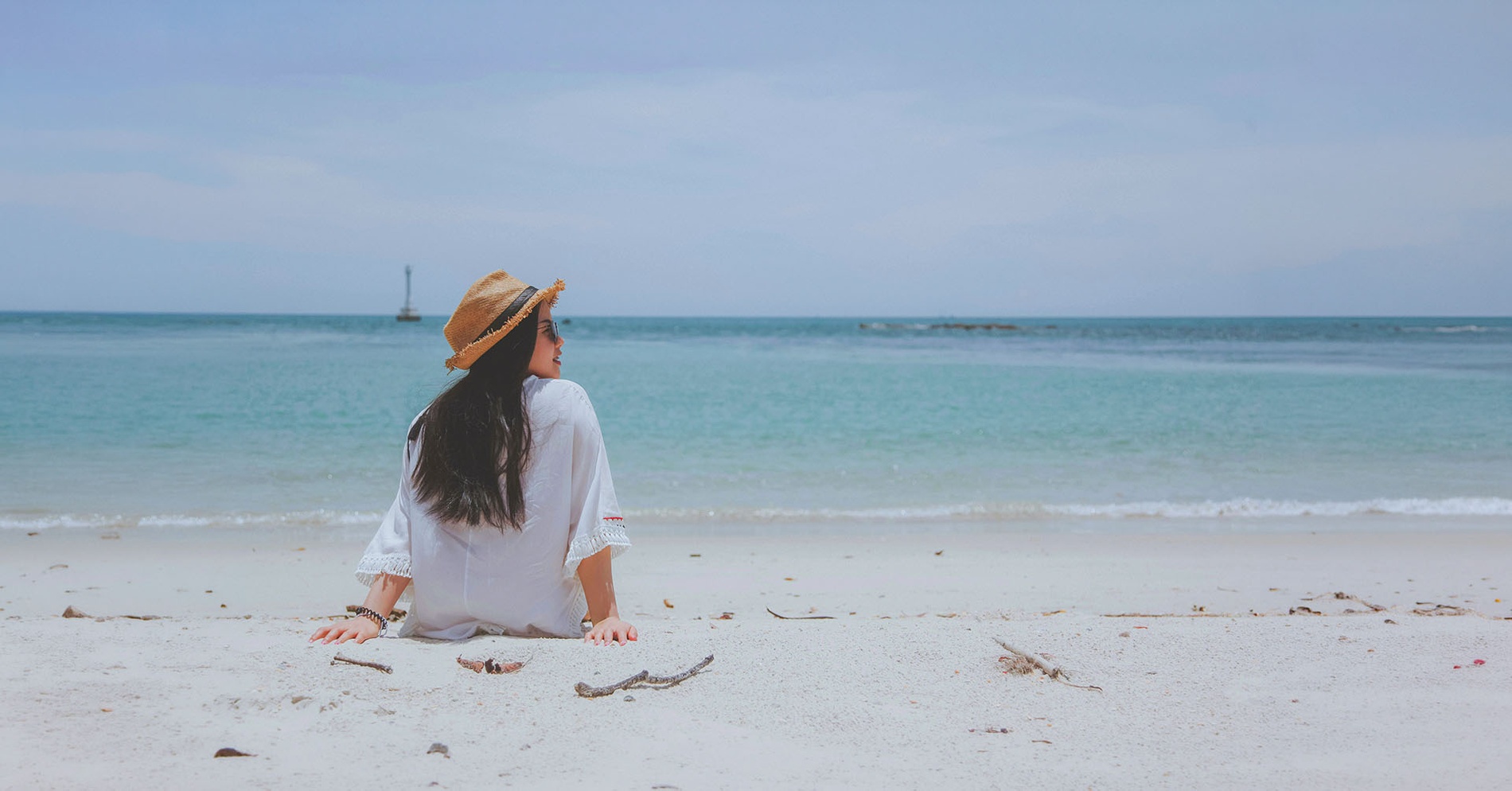 A woman in a straw hat sits on a sandy beach, looking out at the ocean.