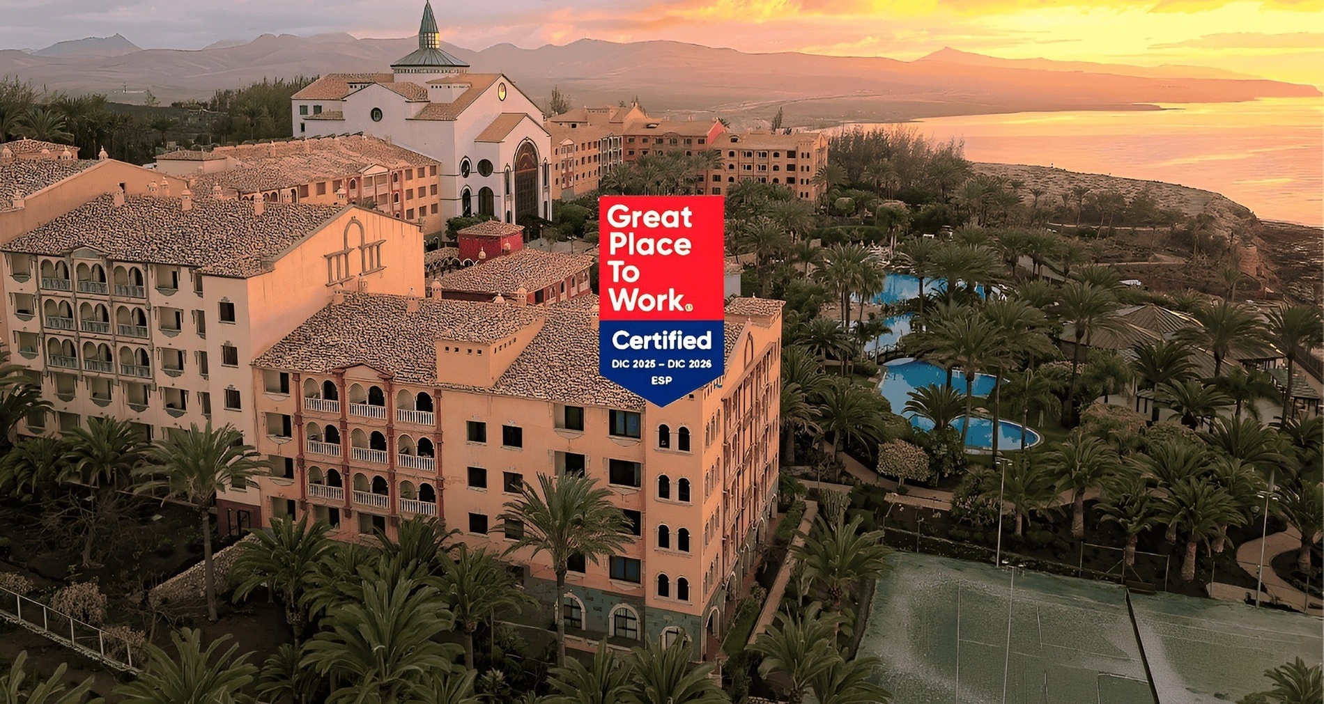 Una vista aerea mostra un complesso di edifici con tetti di tegole e palme, con montagne e il mare sullo sfondo al tramonto, e un logo "Great Place To Work Certified" sovrapposto.
