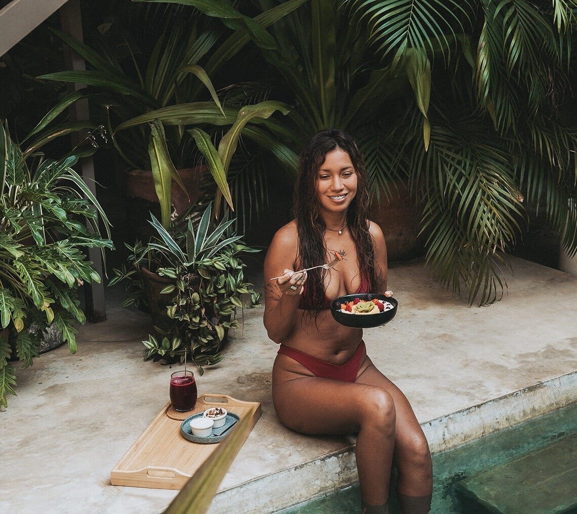 una mujer en bikini está sentada en el borde de una piscina comiendo un tazón de comida