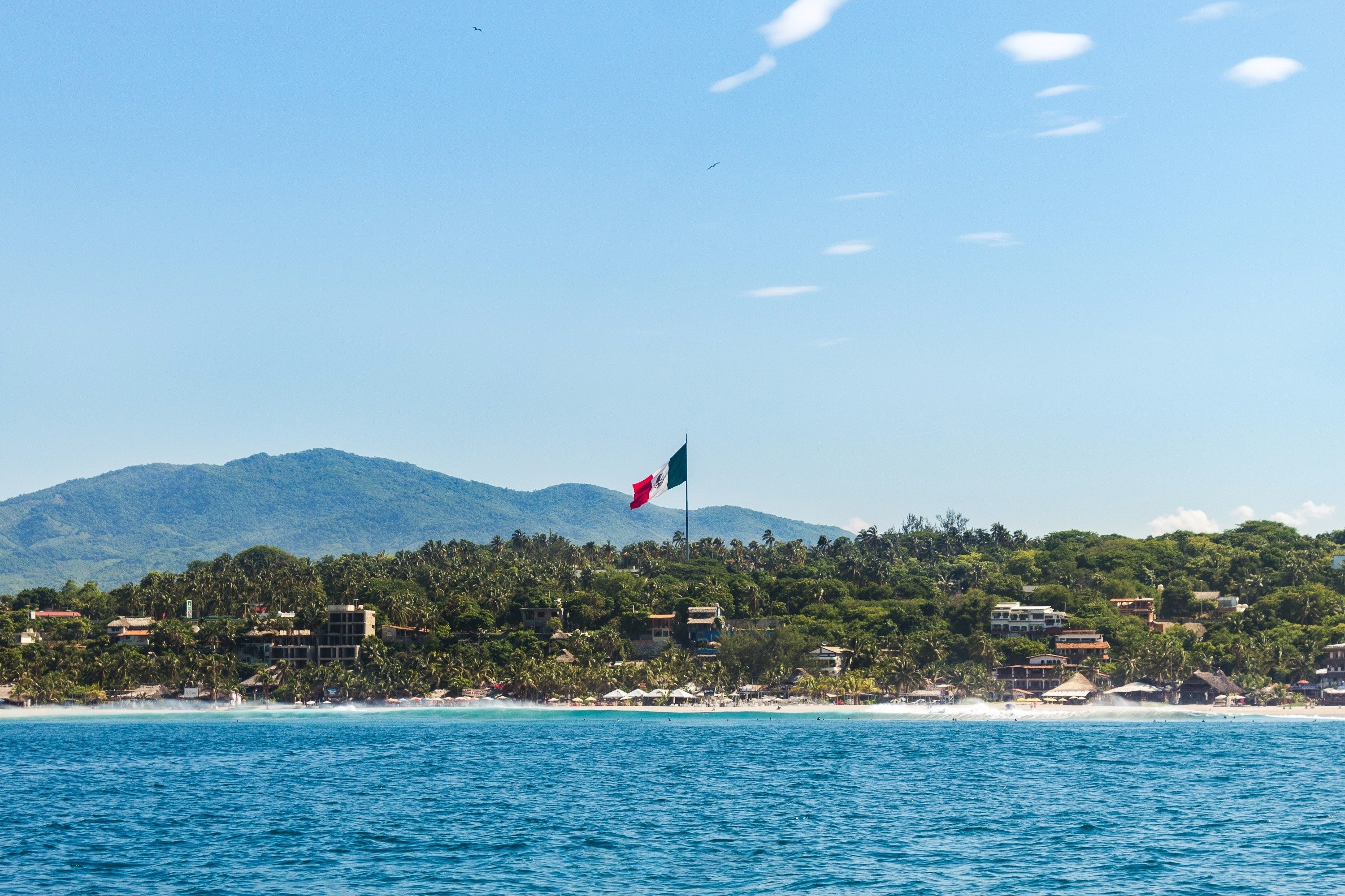 a large mexican flag flies over a beach with mountains in the background