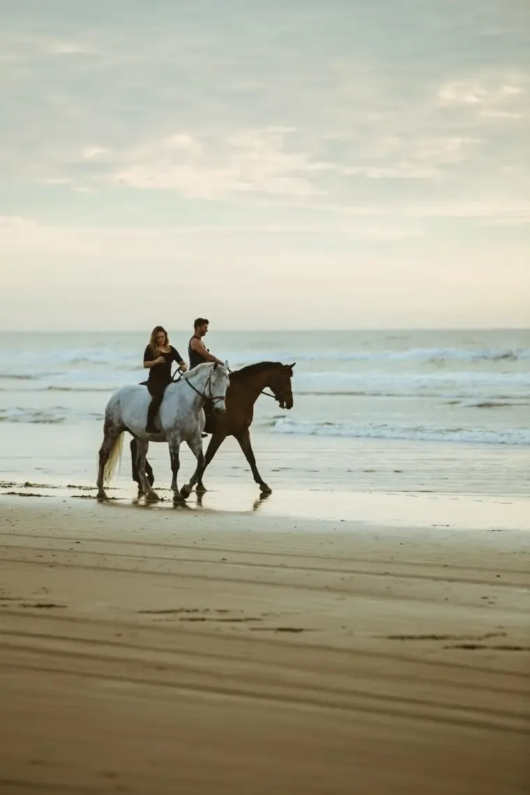 two people riding horses on the beach under a rainbow