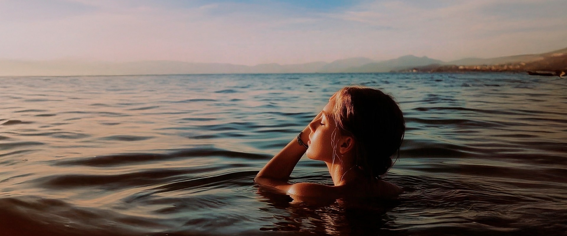 Una mujer se sumerge en el agua con los ojos cerrados y el rostro iluminado por el sol del amanecer o atardecer, con un paisaje montañoso en el fondo.