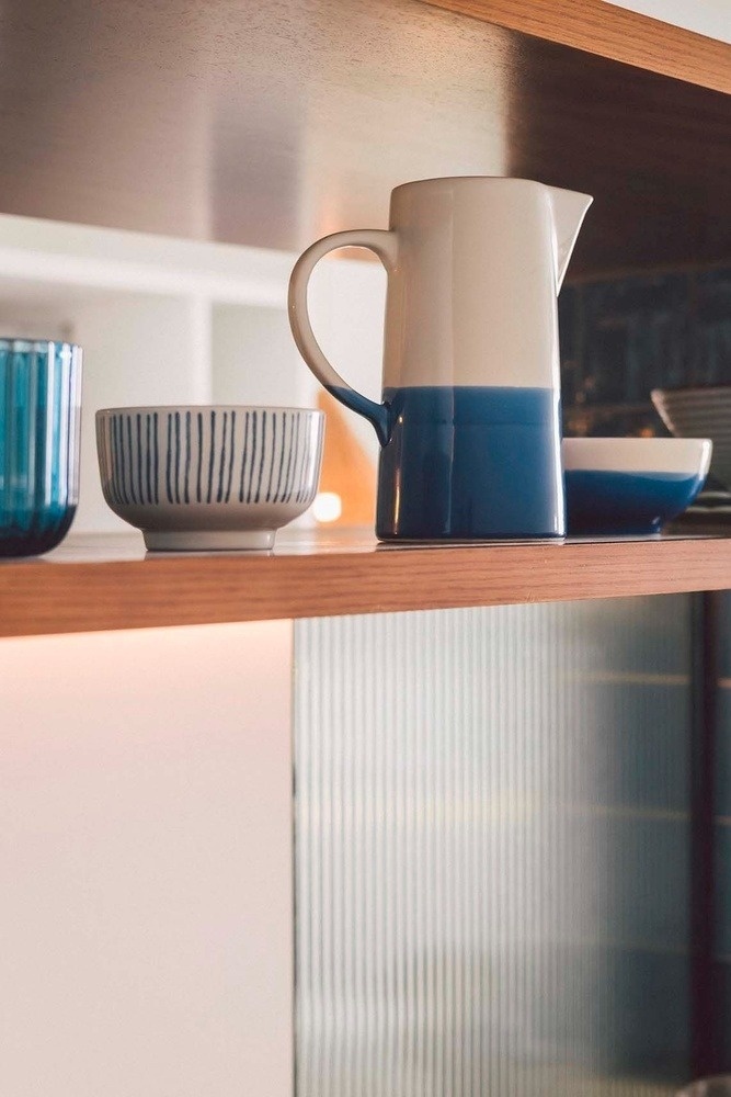A blue and white two-toned pitcher, a striped bowl, and a blue glass are displayed on a wooden shelf, illuminated above a ribbed glass cabinet.