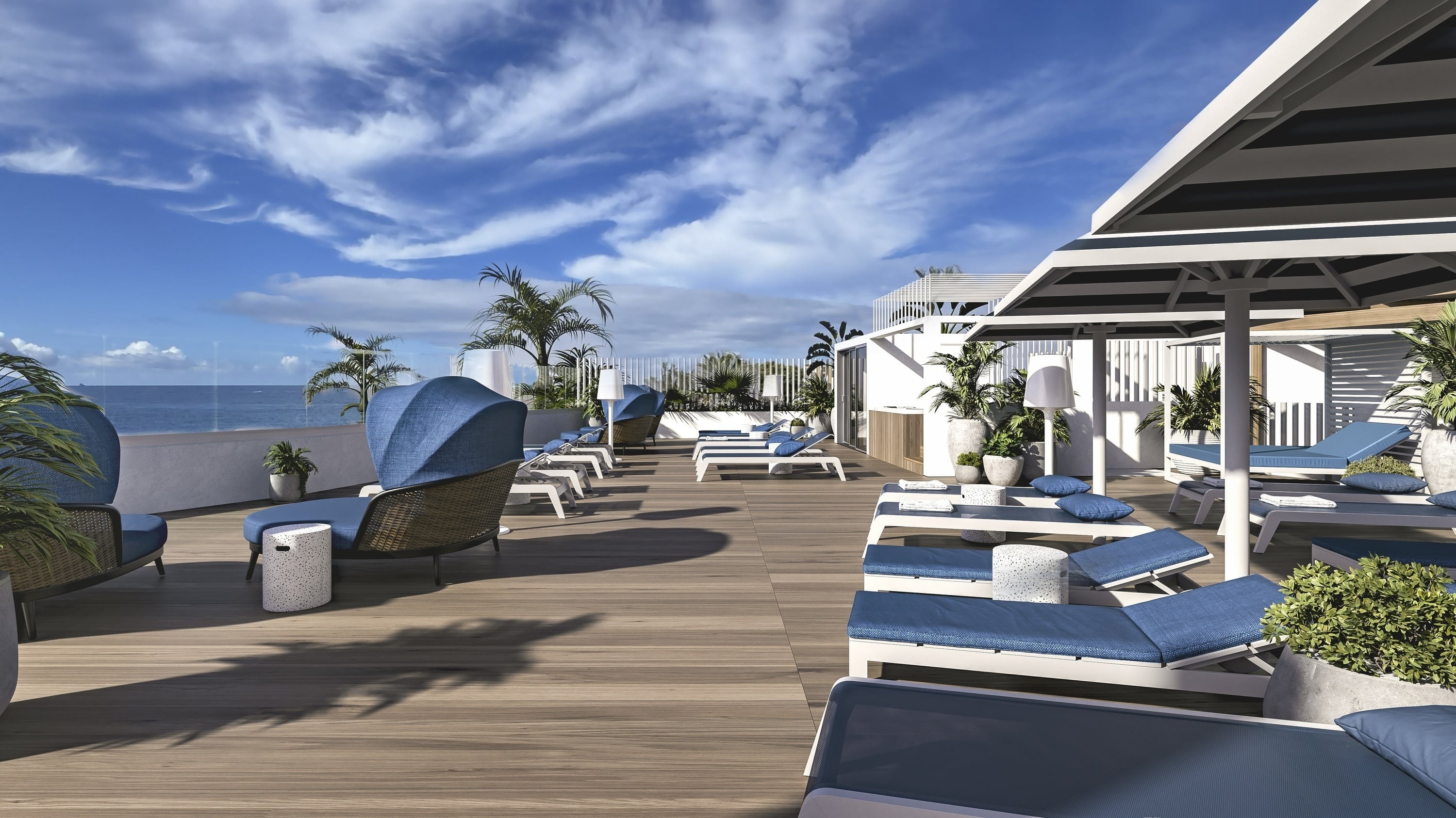 This image captures a luxurious outdoor lounge area on a wooden deck, featuring numerous blue-cushioned sun loungers and umbrellas, with a serene ocean view under a clear blue sky.