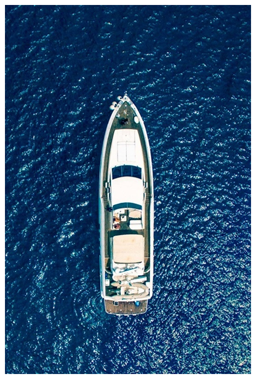 Vista aérea de un moderno yate de lujo de color blanco navegando sobre el mar de un azul profundo. La toma cenital muestra la cubierta superior de la embarcación con zonas de estar y un toldo blanco en la parte trasera. El agua presenta un patrón de pequeñas olas y el sol ilumina la superficie.