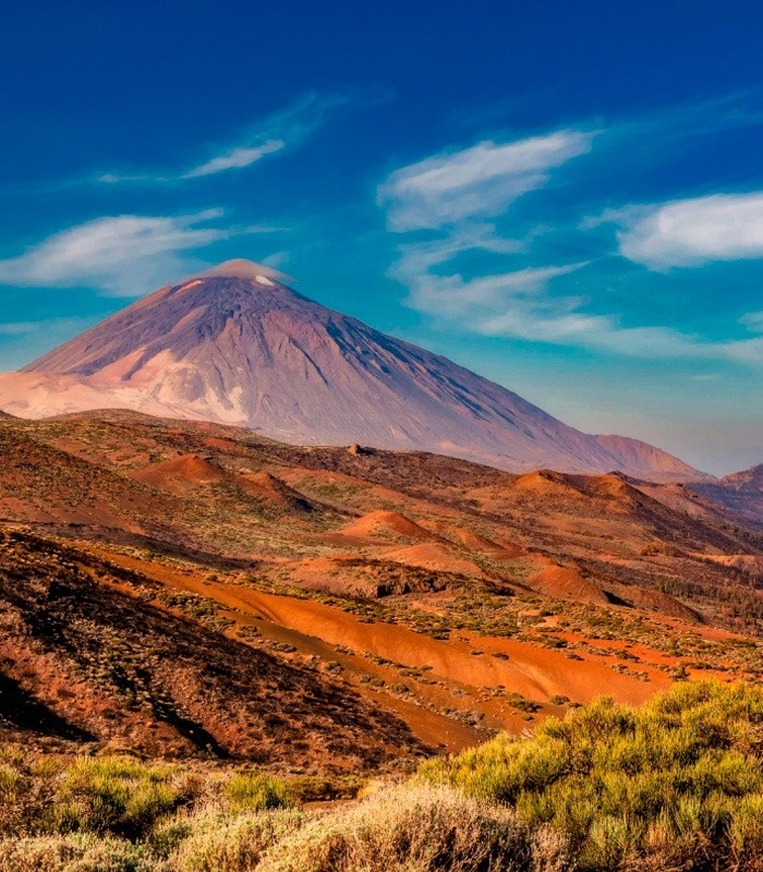 A towering volcanic mountain, capped with a cloud, overlooks a rugged, reddish-brown landscape with sparse vegetation under a vibrant blue sky.