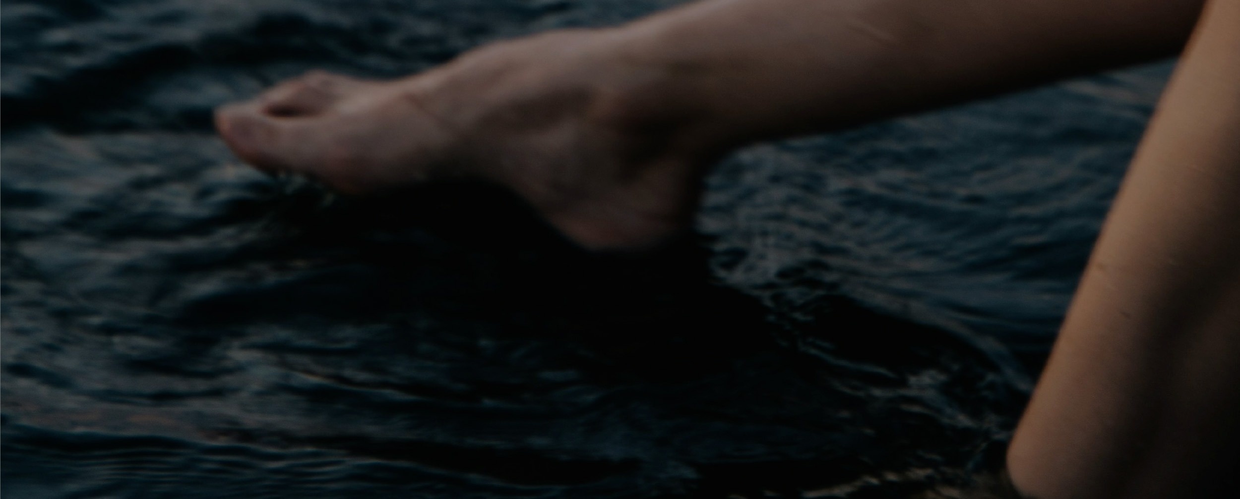 A woman is seen from behind, relaxing in an infinity pool, gazing out at the vast ocean under a clear sky.