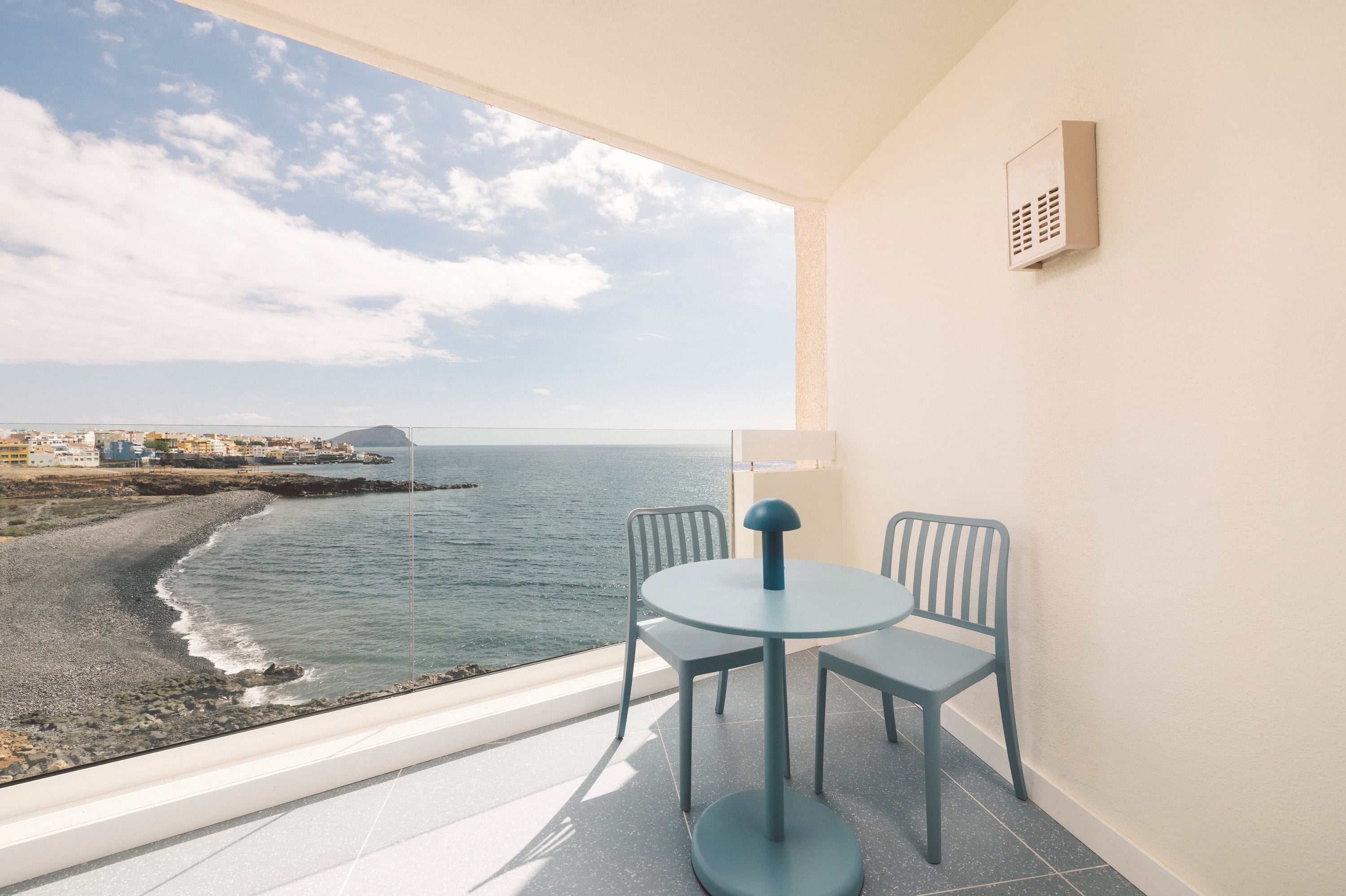 A minimalist balcony with a teal table and chairs overlooks a rocky beach and the ocean, with a distant town under a cloudy sky.