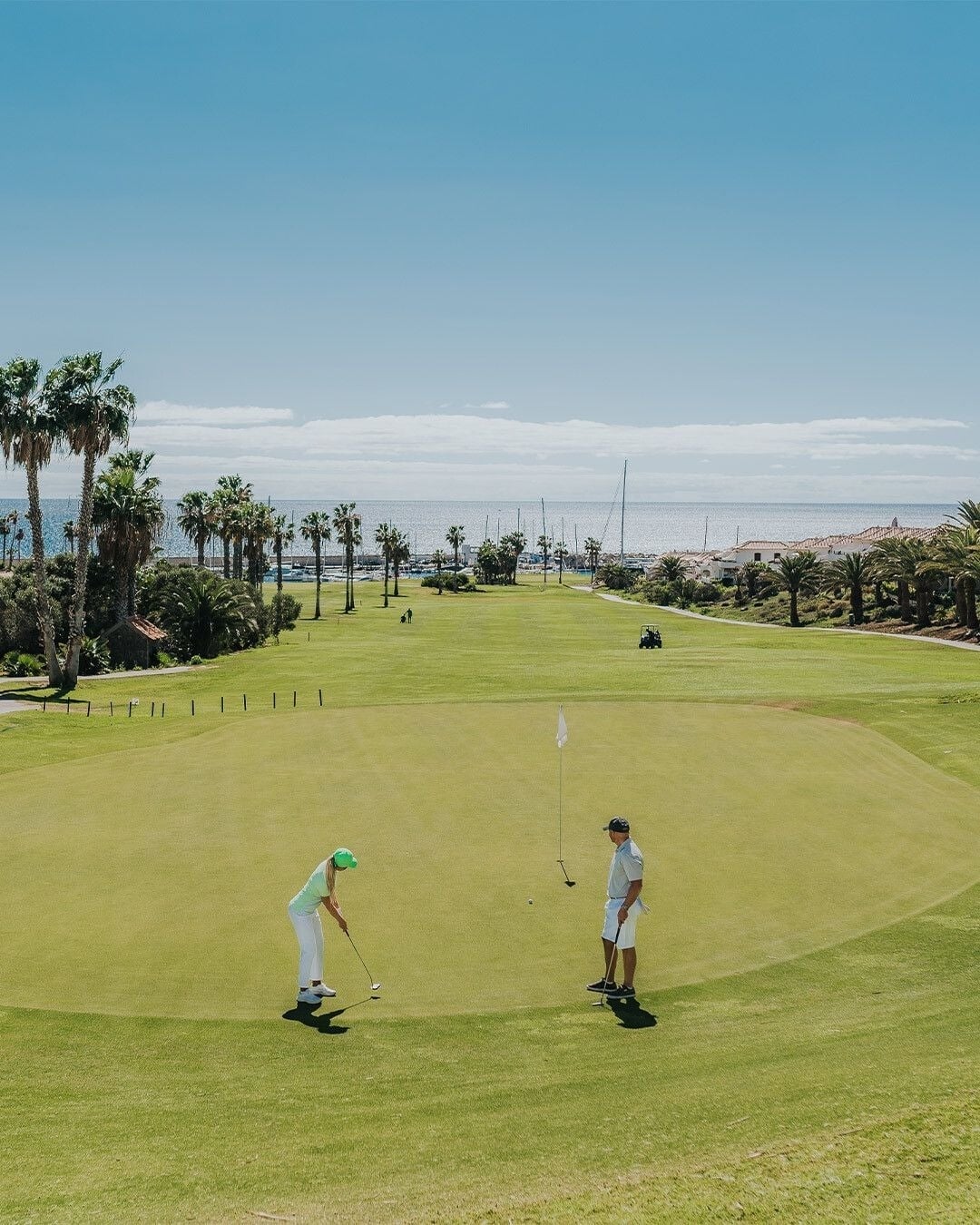 Campo de golf verde con vistas al océano en un día soleado, rodeado de palmeras y vegetación costera. En primer plano, una mujer con gorra verde y ropa blanca está a punto de dar un golpe en el green, mientras un hombre la observa cerca de la bandera. Al fondo se distingue una pequeña marina y edificios de apartamentos.