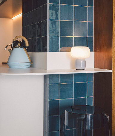 En un ambiente moderno, una tetera azul claro y una lámpara iluminada reposan sobre una repisa blanca, frente a una pared de azulejos azules cuadrados.