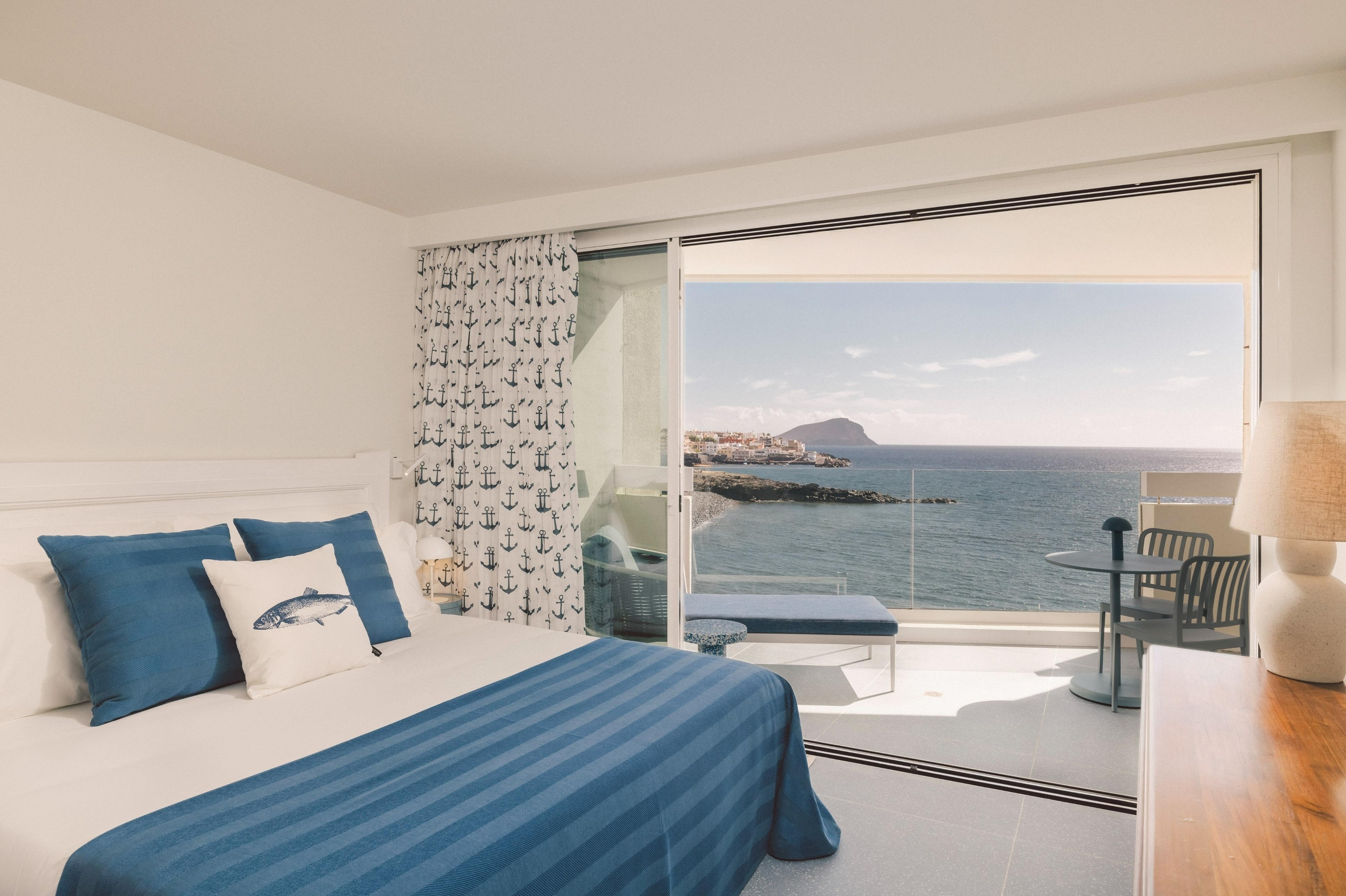 A brightly lit bedroom with a nautical theme, featuring a bed with blue and white linens, opens onto a balcony overlooking the sea and a distant coastal town.