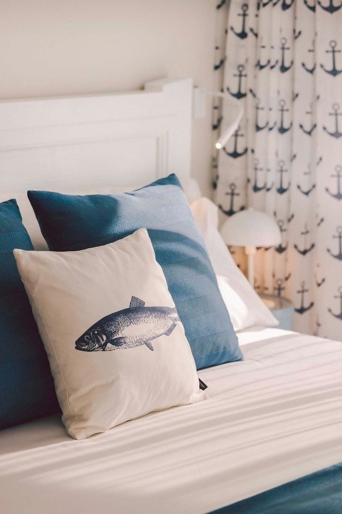 A bright bedroom features a bed adorned with blue and fish-printed pillows, white sheets, and anchor-patterned curtains in the background.