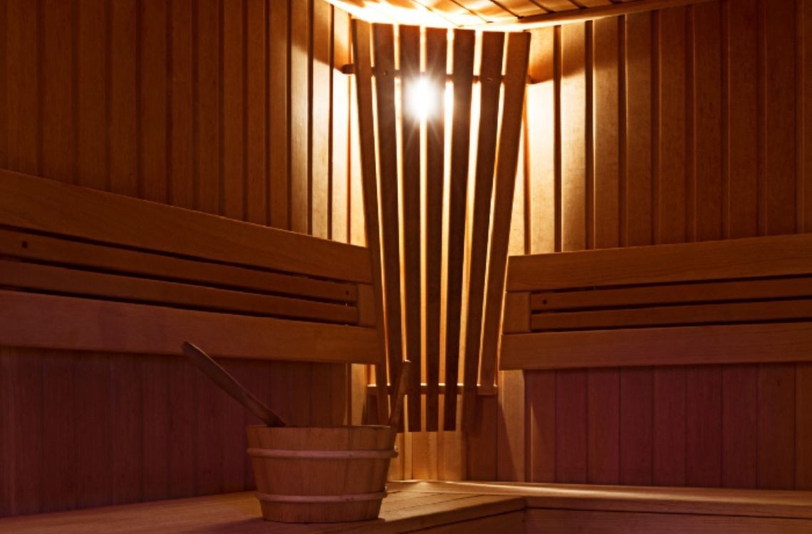 El interior de una sauna de madera con una luz brillante filtrándose a través de listones verticales y un cubo con cucharón en un banco.