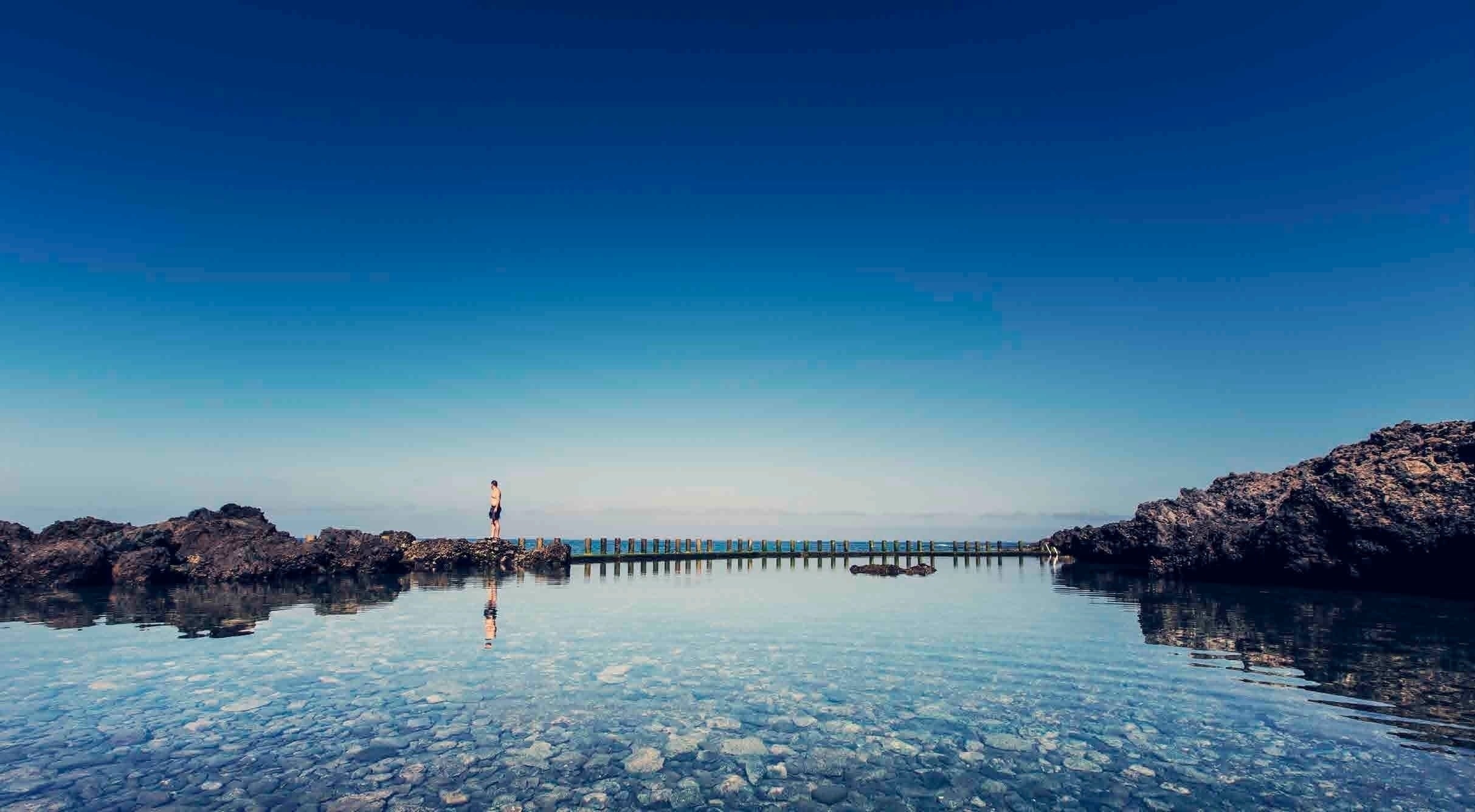 Vista aérea de una bahía rocosa con varios catamaranes en aguas cristalinas, flanqueada por montañas áridas bajo un cielo azul.