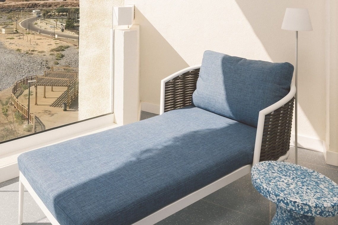 A modern blue chaise lounge with white and wicker accents sits on a sunlit balcony next to a window overlooking a dry, desert-like landscape, accompanied by a small blue patterned table and a floor lamp.