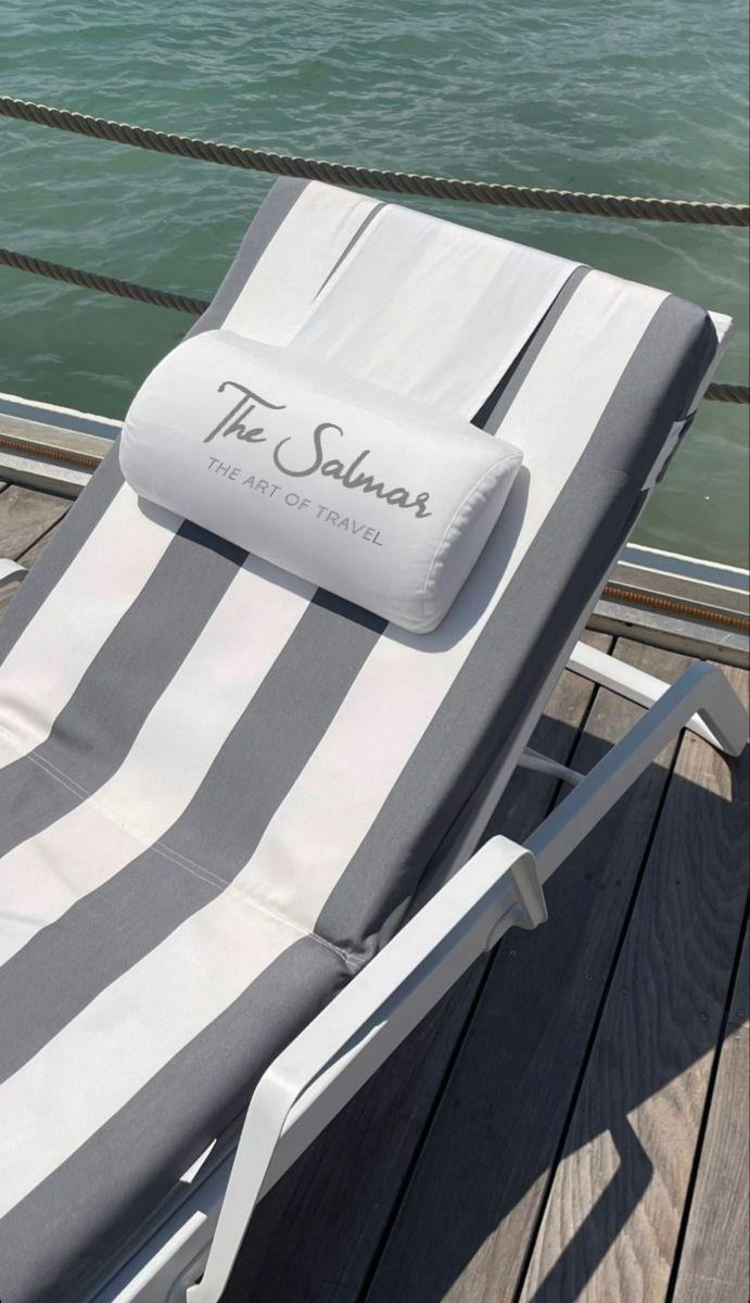 A grey and white striped lounge chair with a branded pillow rests on a wooden pier overlooking calm blue-green water, secured by a rope railing.