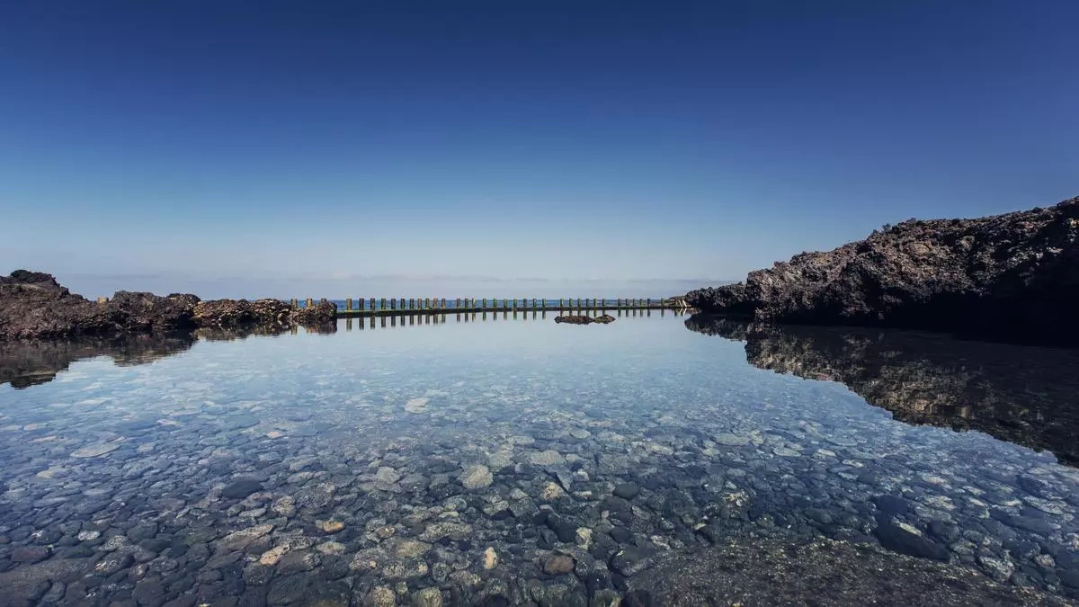 Un hombre se sitúa sobre rocas al borde de una tranquila piscina natural de agua cristalina con el fondo rocoso visible, bajo un vasto cielo azul, reflejándose en la calma superficie.