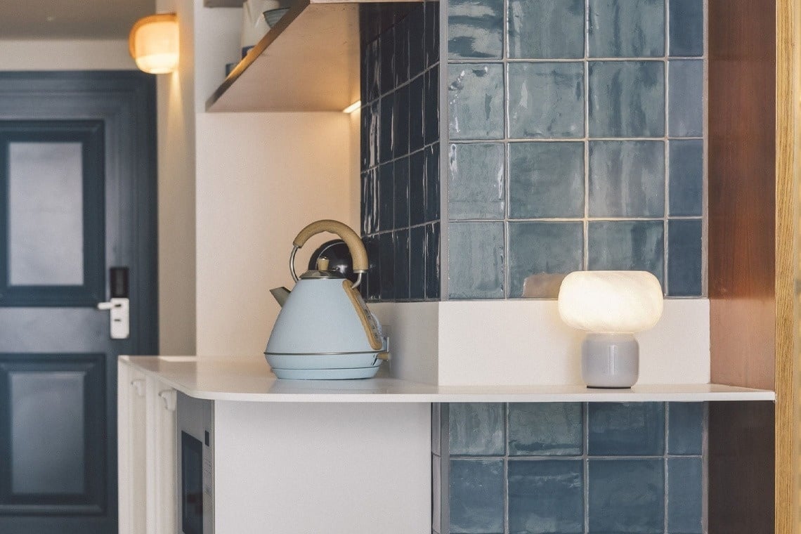 A light blue kettle and a glowing white lamp are placed on a white counter, set against a backdrop of glossy blue-green square tiles, with a dark blue door and a wall sconce visible in the background.