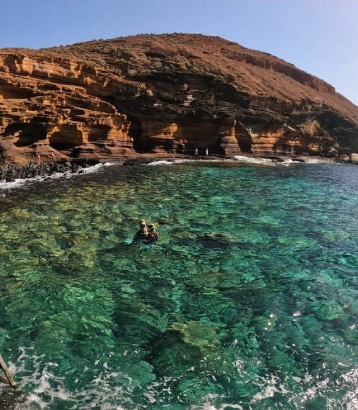People are snorkeling in incredibly clear turquoise water alongside a large, layered rocky cliff with figures standing on its base.