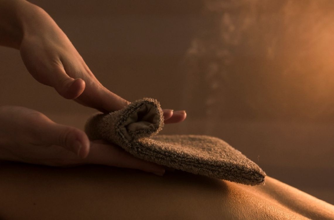 En una escena con iluminación cálida, unas manos sostienen suavemente una toalla sobre una espalda.