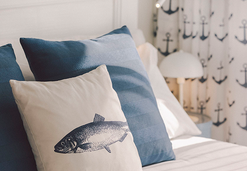 Una cama decorada con cojines azules y uno con un estampado de pez, y cortinas de anclas al fondo, presenta una temática náutica.
