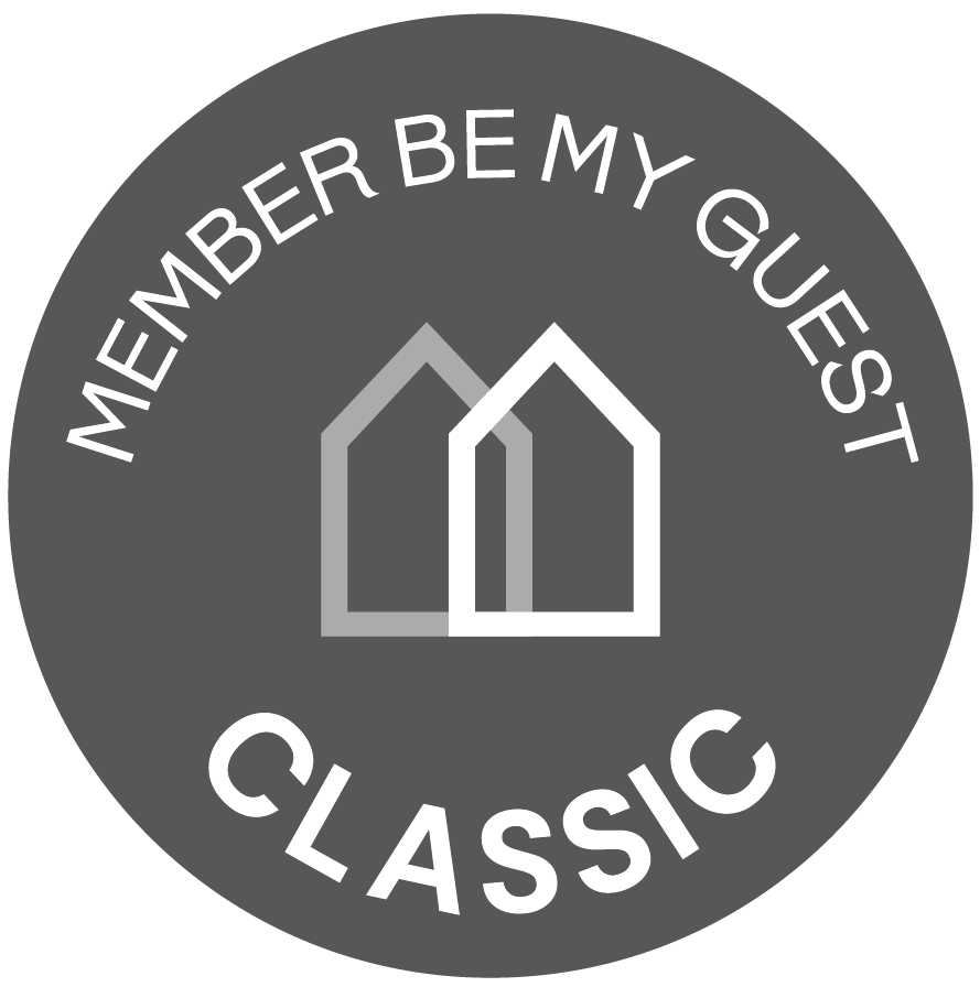 El logo muestra un círculo gris oscuro con dos siluetas de casas superpuestas en el centro, y el texto blanco "MEMBER BE MY GUEST" curvado en la parte superior y "CLASSIC" en la inferior.