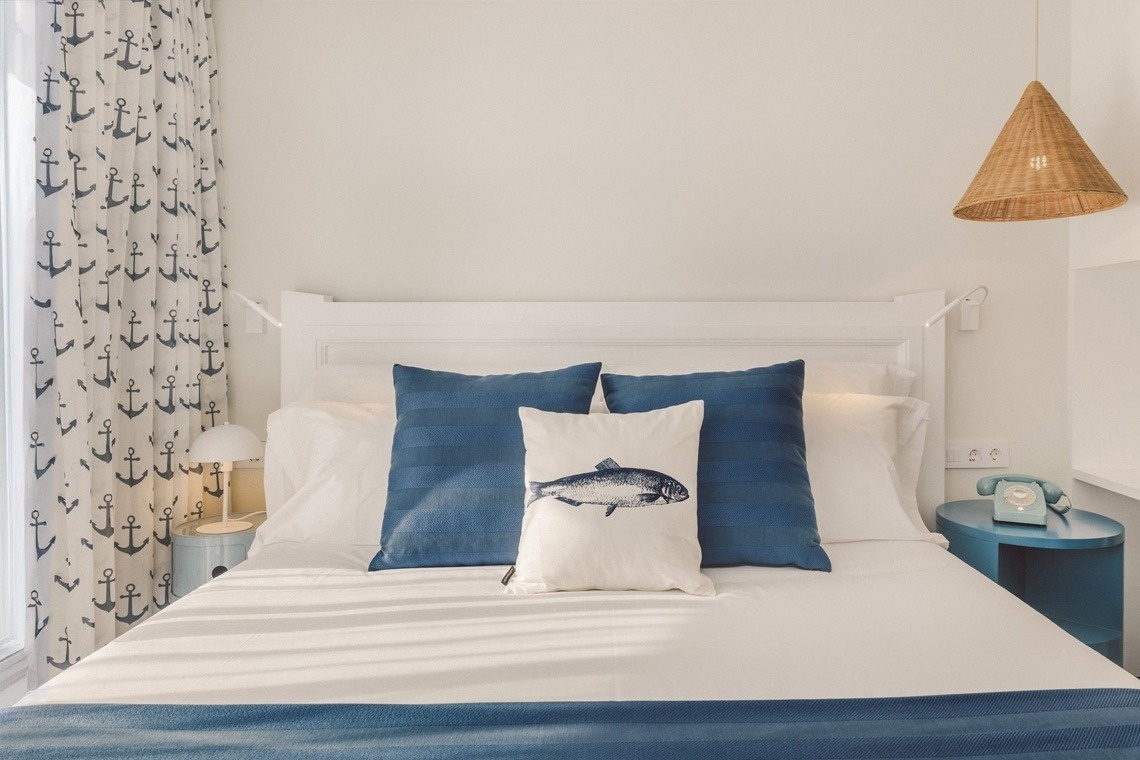 A brightly lit bedroom features a nautical theme with anchor-patterned curtains, white and blue bedding, a fish-print pillow, and a light blue nightstand with a retro telephone.