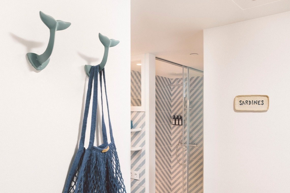 Two whale-tail shaped hooks, one holding a blue mesh bag, adorn a white wall next to a shower with chevron tiles and a 