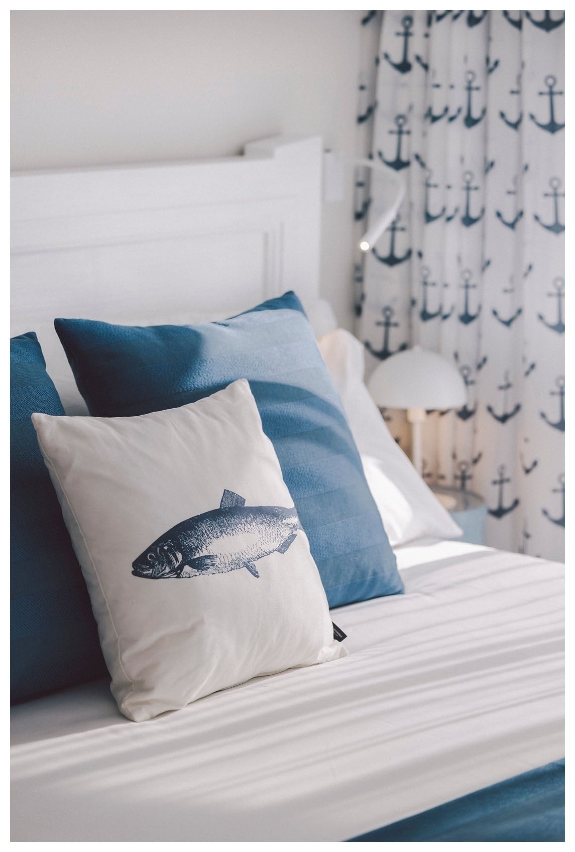 Detalle de cama en habitación de The Salmar Tenerife con decoración náutica, cojín con ilustración de pez, textiles azules y cortinas con motivos de anclas.
