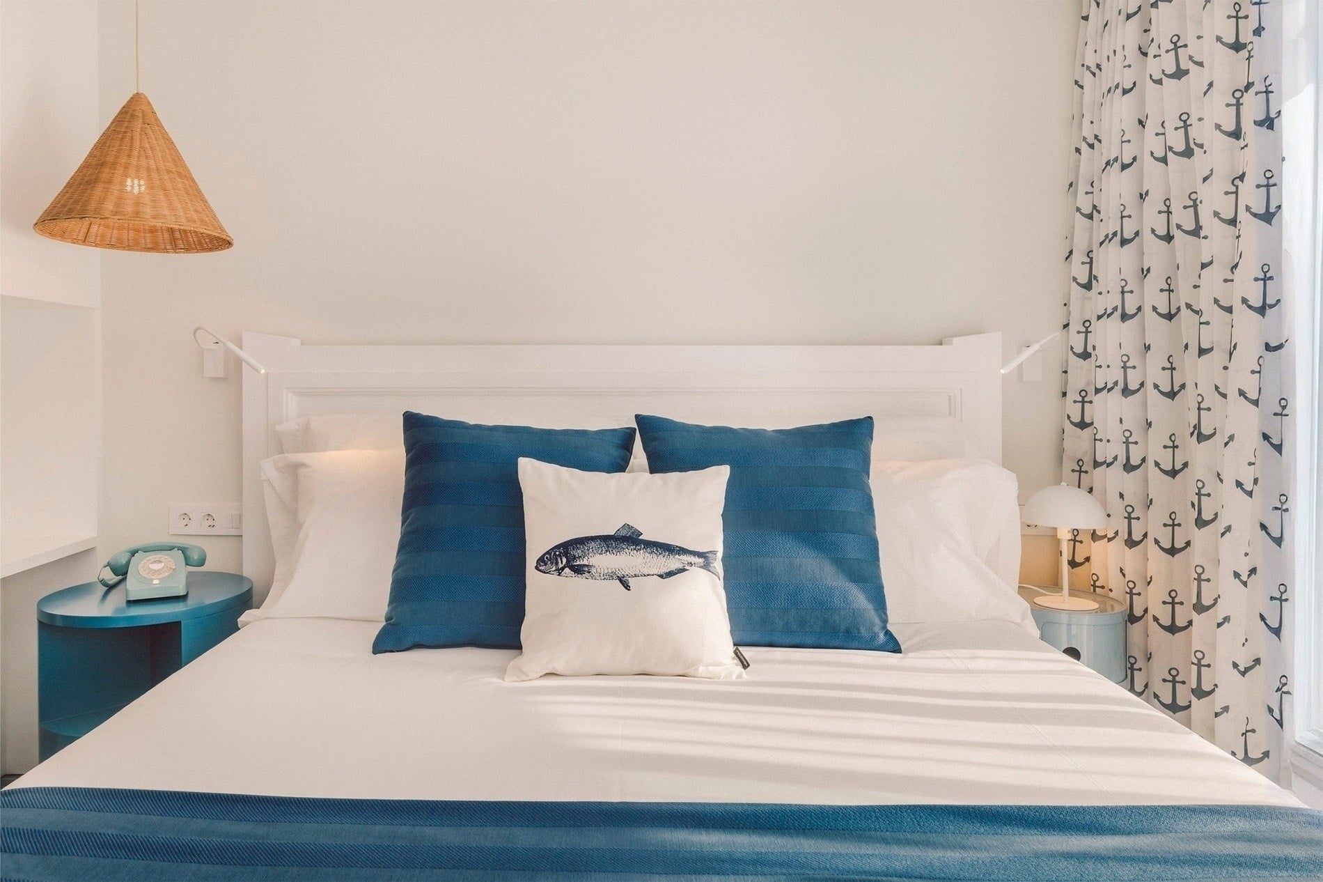 Un dormitorio de estilo náutico presenta una cama con ropa de cama blanca y azul, cojines con motivos marinos y cortinas estampadas con anclas, bajo una lámpara colgante de mimbre.