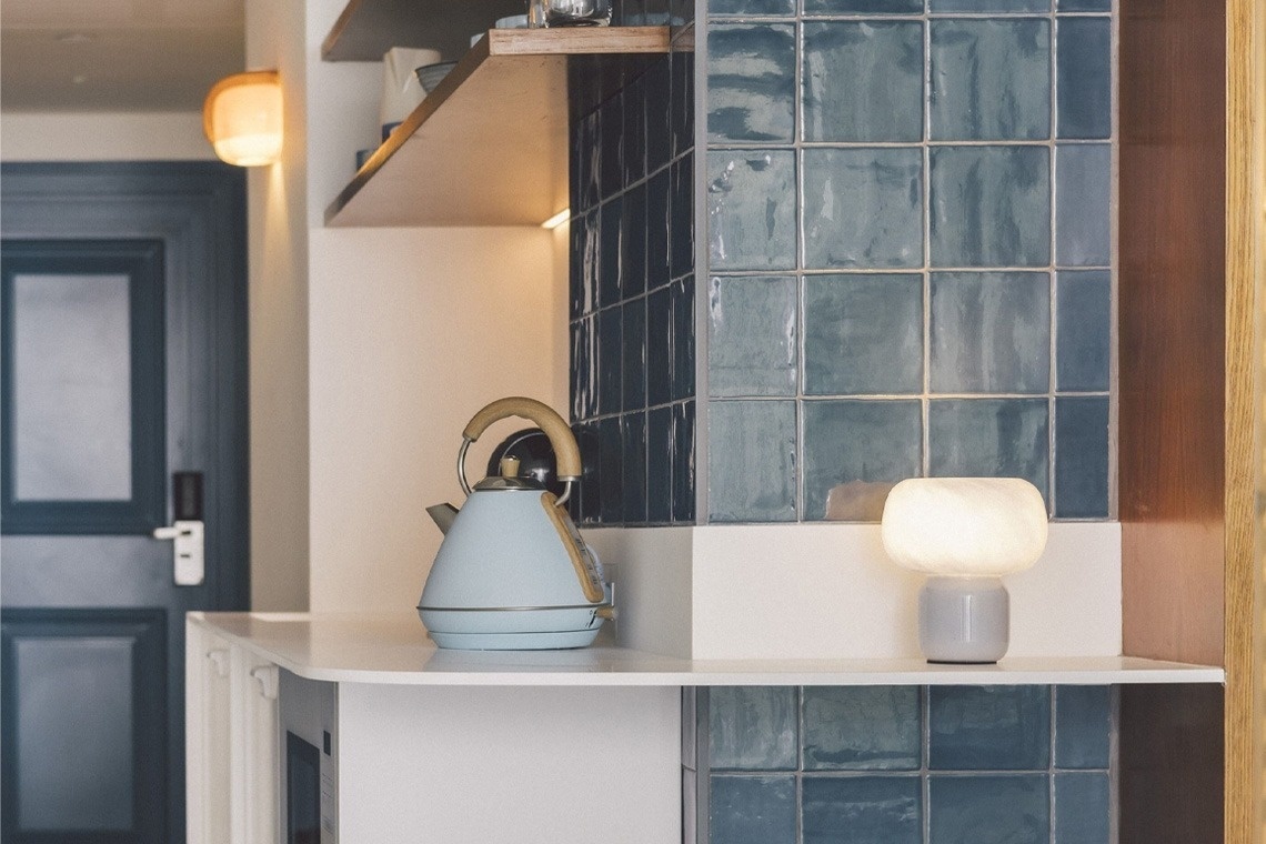 A light blue kettle and a glowing white lamp sit on a clean white countertop, flanked by blue tiled walls and wooden shelving, with a dark blue door visible in the background.