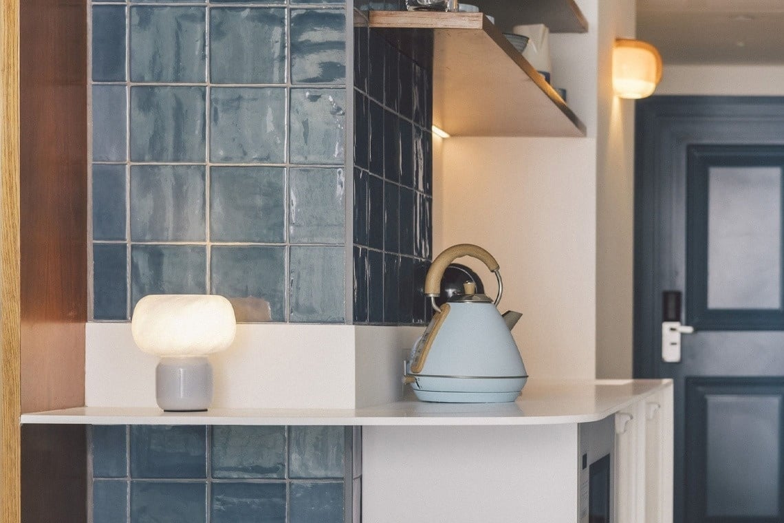 A light blue kettle and a glowing white lamp are placed on a white counter in a kitchen area featuring blue tiled walls and a dark door in the background.