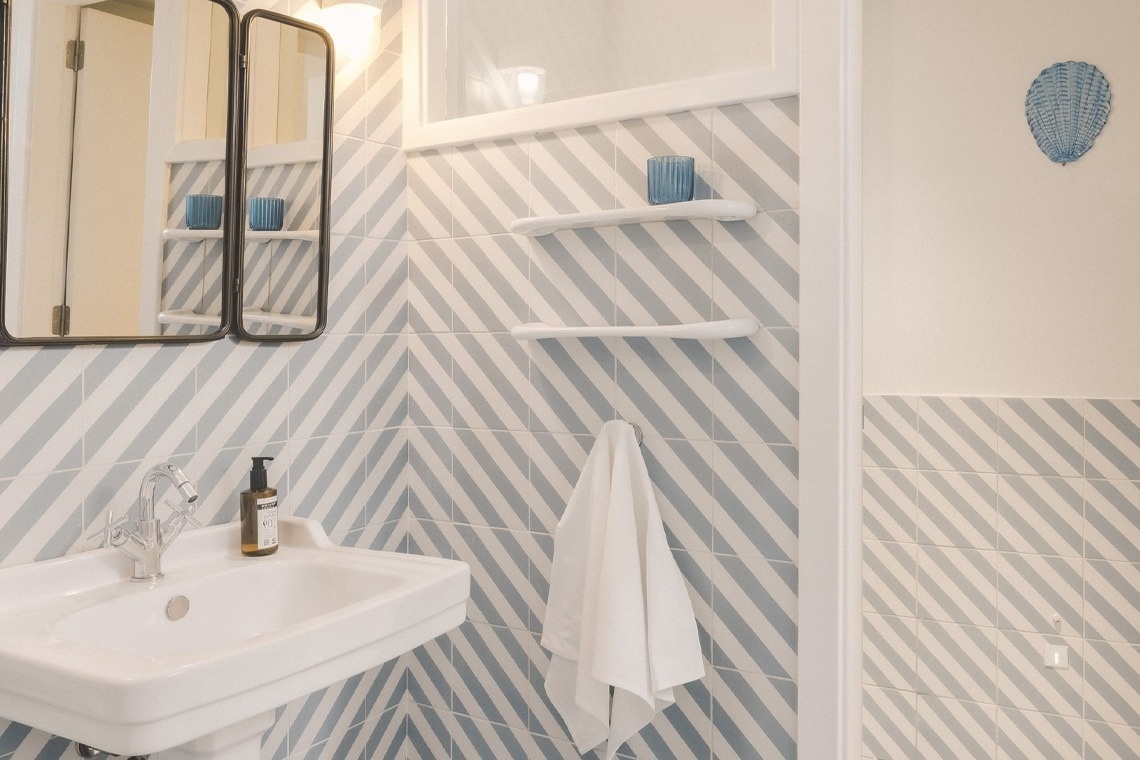 A modern bathroom features a white sink and mirror set against a wall with striking blue and white diagonally striped tiles, accented by white shelves, a towel, and a decorative blue seashell.
