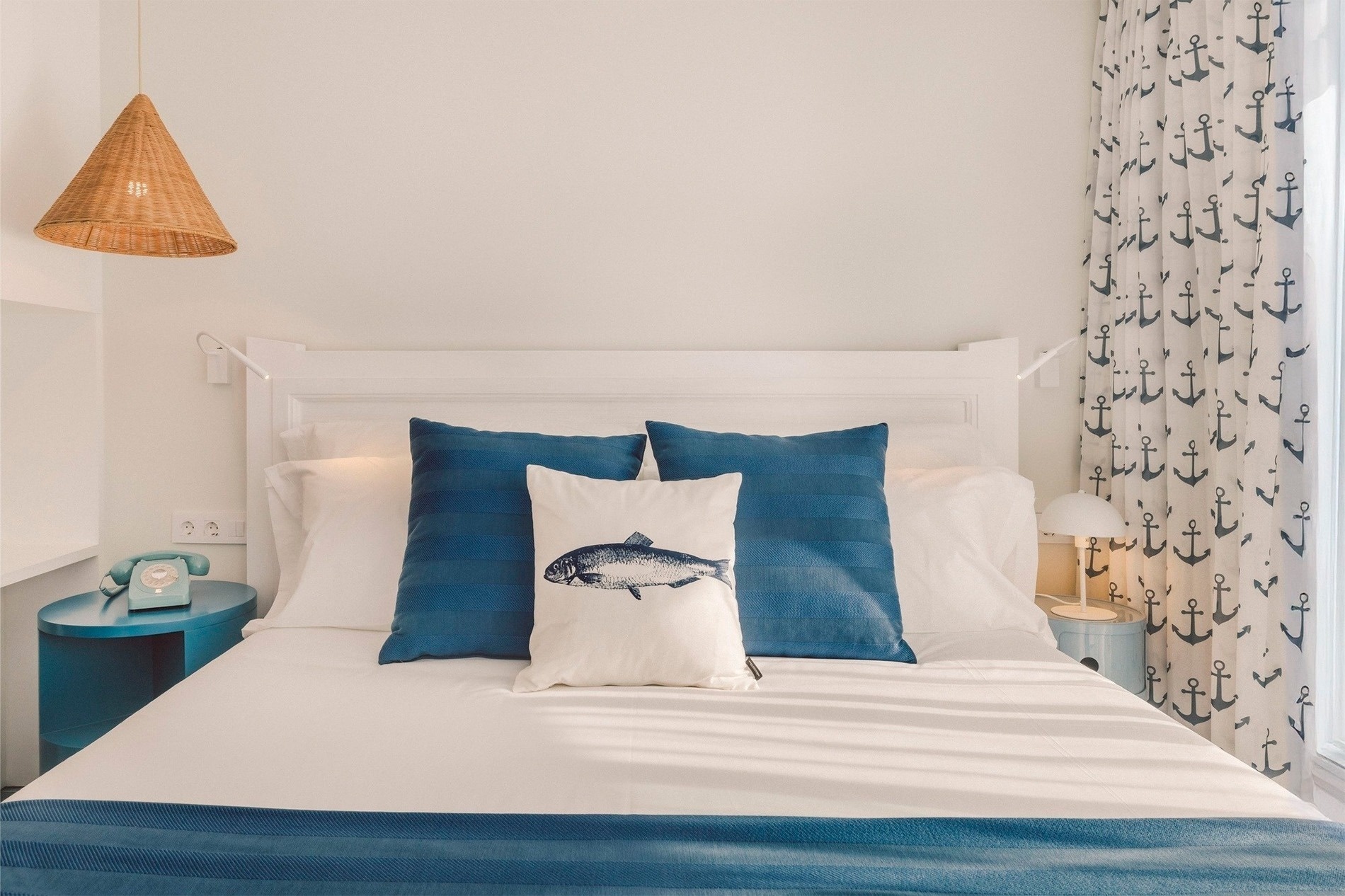Una luminosa habitación de estilo náutico presenta una cama con sábanas blancas y detalles en azul, almohadas con un diseño de pez y cortinas estampadas con anclas.