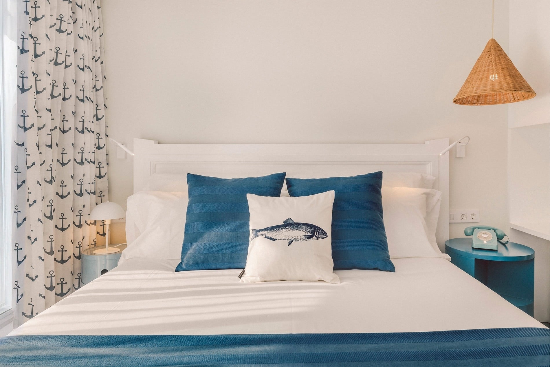 A nautical-themed bedroom features a white bed with blue and white bedding, a fish-printed pillow, anchor-patterned curtains, and blue bedside tables, one with a vintage telephone.