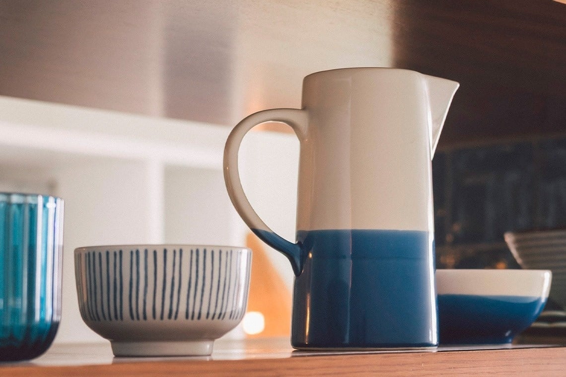 A white and deep blue two-toned ceramic pitcher stands prominently on a wooden shelf, accompanied by a white and blue striped bowl, a blue glass, and another blue-dipped bowl in the blurred background.