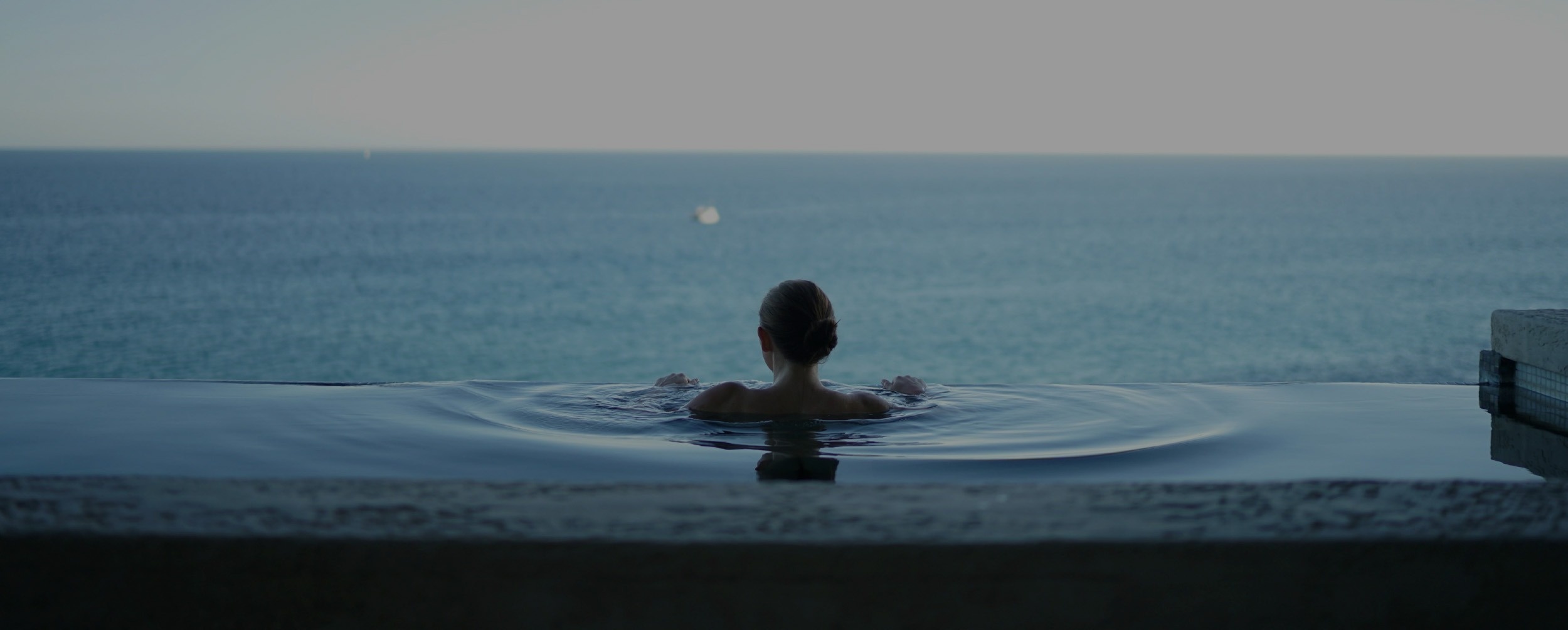 Una mujer, vista de espaldas, se encuentra en una piscina infinita mirando hacia el océano.