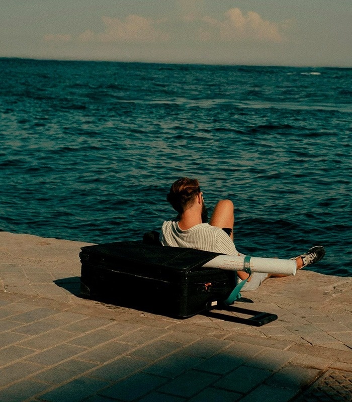 A man with his back to the viewer sits leaning against a black suitcase on a paved dock, gazing out at the expansive blue sea.
