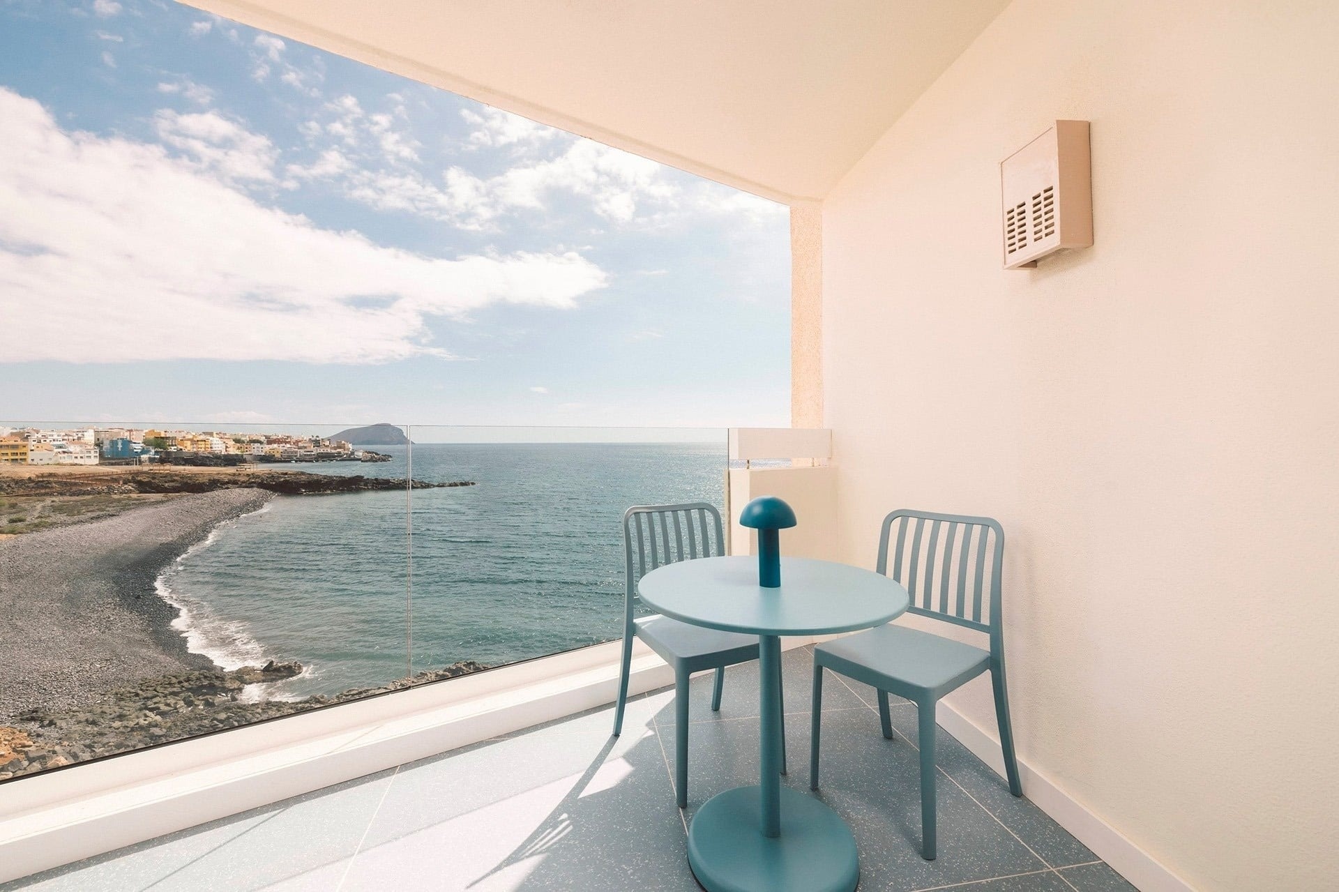 A bright balcony with turquoise furniture offers a panoramic view of a rocky beach, a coastal town, and the blue ocean under a partly cloudy sky.