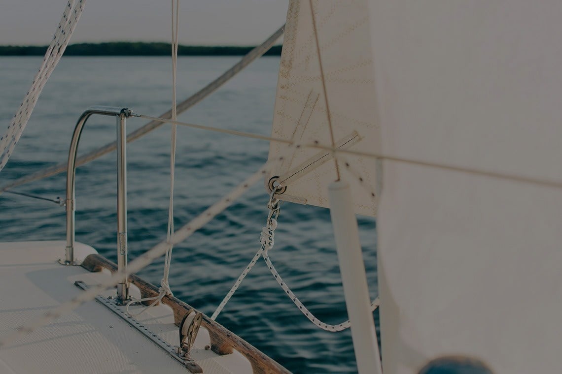 A close-up view from a sailboat shows part of the deck, rigging, and a sail against a background of calm blue water.