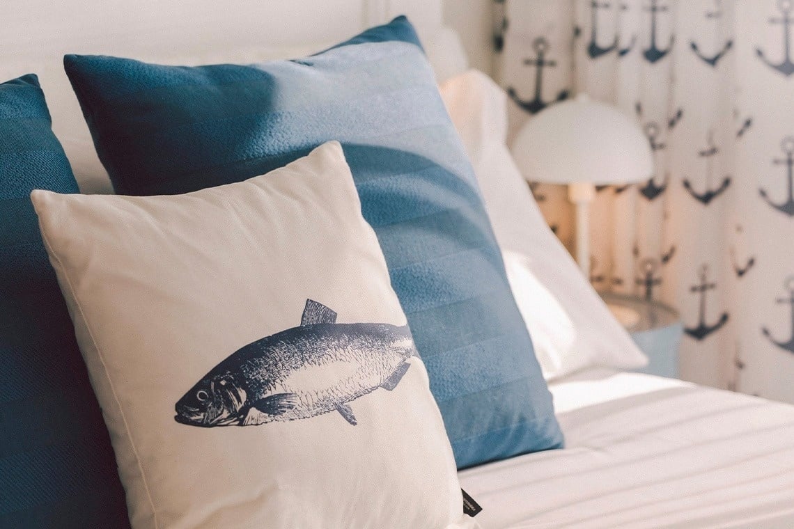 Nautical-themed bedding with blue and off-white pillows, one featuring a fish illustration, is shown against a soft-focused background of anchor-patterned curtains.