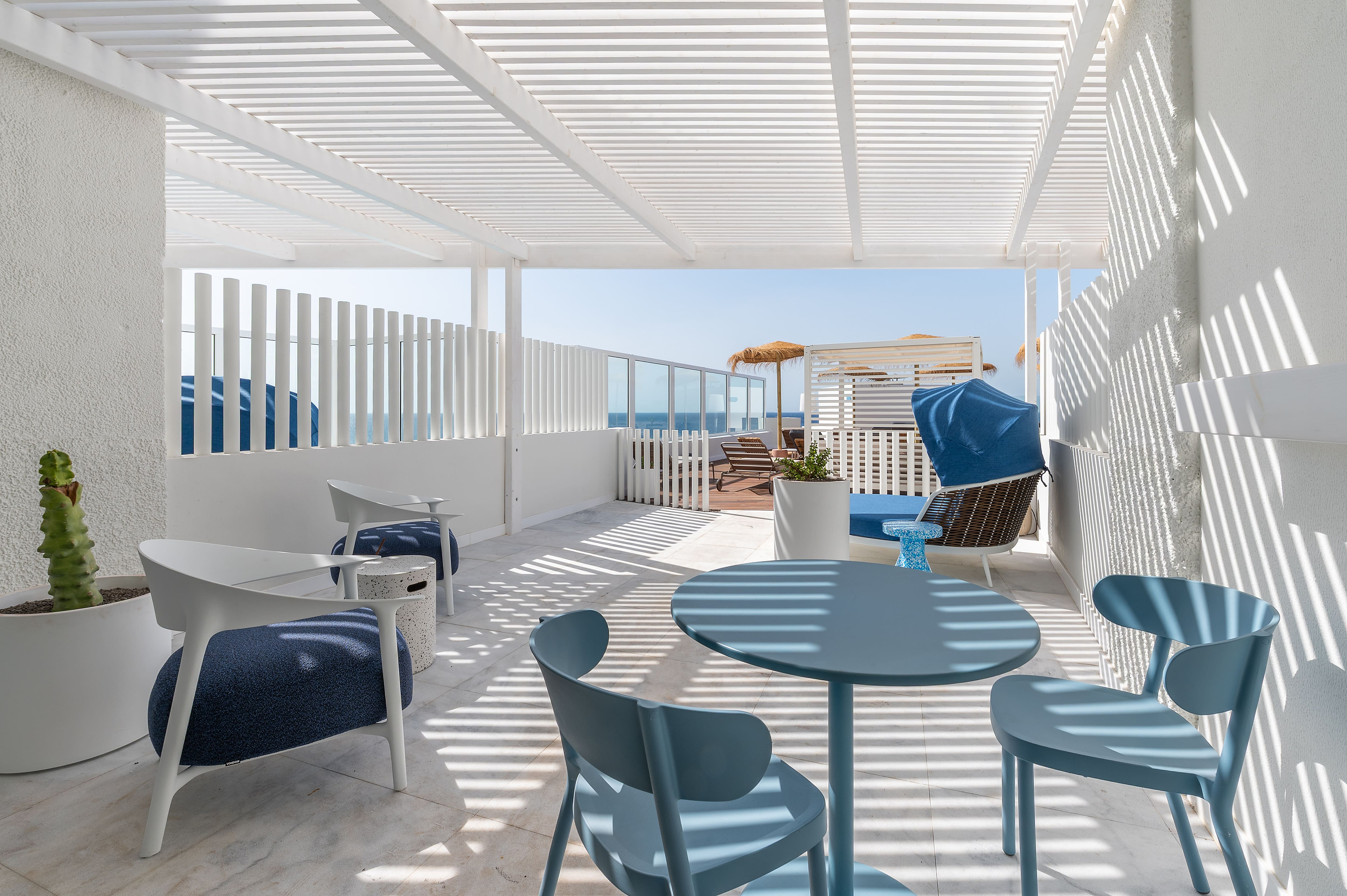 Terraza de hotel con sillas y mesa azul, vistas al mar y pérgola blanca con sombra rayada Satocan: The Salmar.