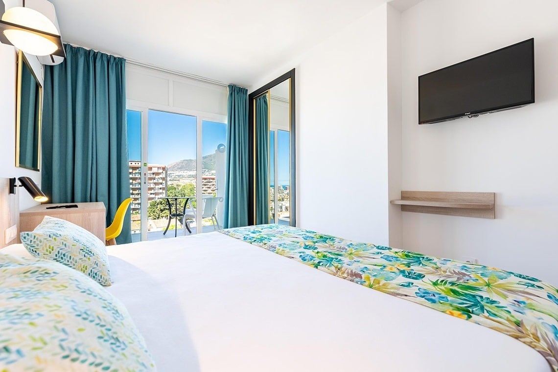 Habitación de hotel moderna con cama doble, balcón, TV y vistas a la ciudad y montaña.