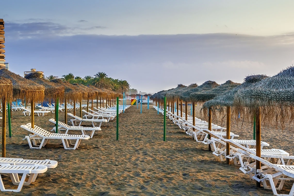 Playa de hotel con filas de sombrillas de paja y tumbonas blancas en la arena para vacaciones.