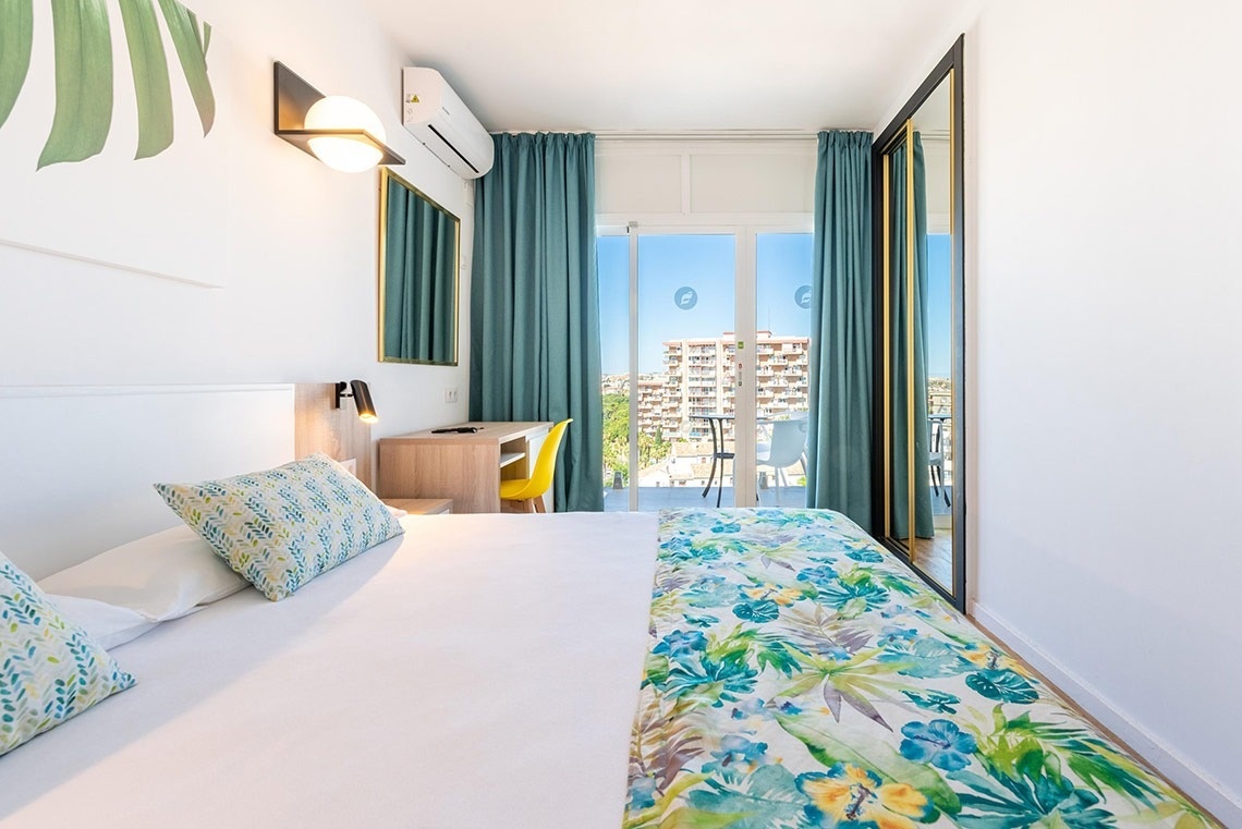 Habitación de hotel luminosa con cama cómoda, balcón, vistas urbanas y diseño fresco tropical.