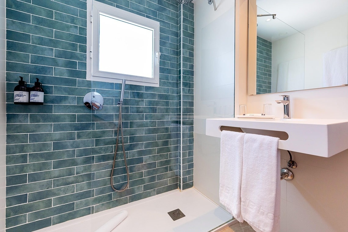 Baño moderno de hotel con ducha de azulejos azules, ventana, lavabo blanco y toallas limpias.