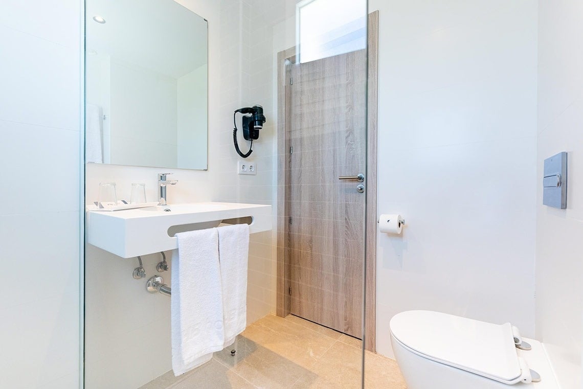 Baño moderno de hotel con lavabo blanco, espejo, secador y WC.